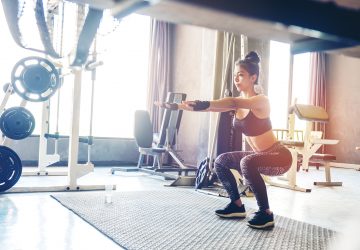 Deep squat of young woman in sportswear doing squat while standing in front of glass at gym