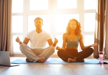 Couple doing yoga exercises at home. They sit on the floor on yoga mats in a lotus position. In front of them is a gray laptop. They smile at each other.